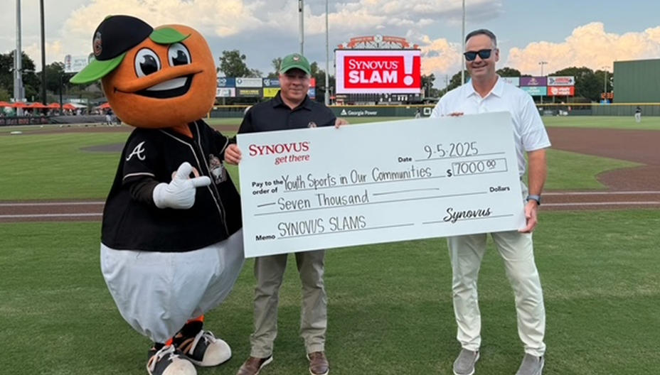 Columbus Clingstones mascot Fuzzy, who has a peach for a head, helps two men hold up a large check for seven thousand dollars representing donations raised for Synovus Slams. The bank donated one thousand dollars for each grand slam home run by the Clingstones in their inaugural season at Synovus Park.